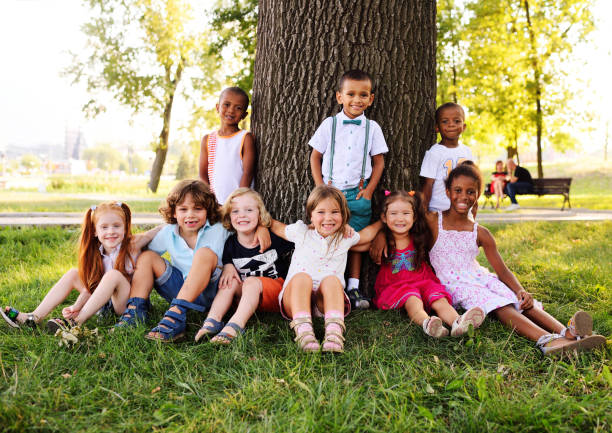 un gruppo di bambini piccoli in abiti colorati che si abbracciano seduti sull'erba sotto un albero in un parco ridendo e sorridendo - preschooler portrait family outdoors foto e immagini stock