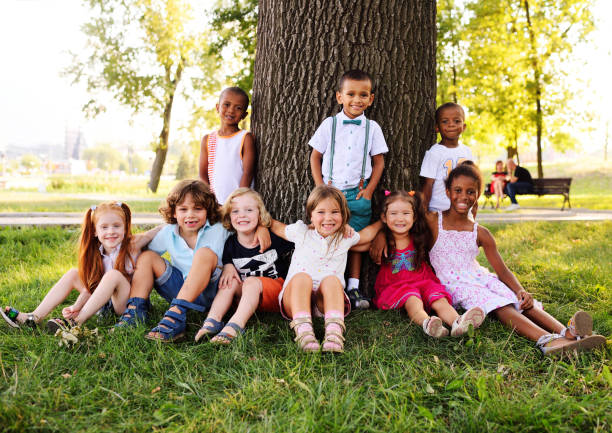 a group of small children in colorful clothes embracing sitting on the grass under a tree in a park laughing and smiling stock photo