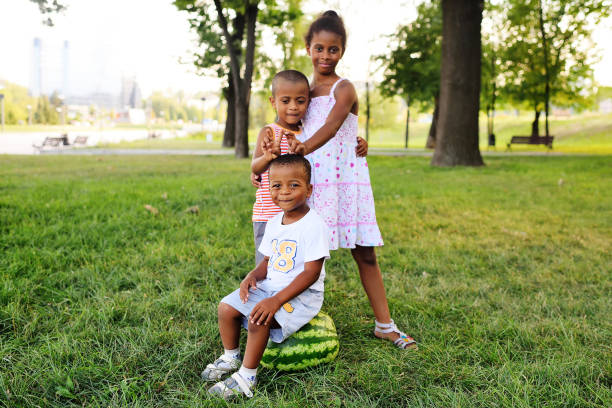 glückliche schwarz-afroamerikanische kinder, die mit einer großen wassermelone im park auf dem rasen spielen und lächeln - sibling baby three people baby girls stock-fotos und bilder