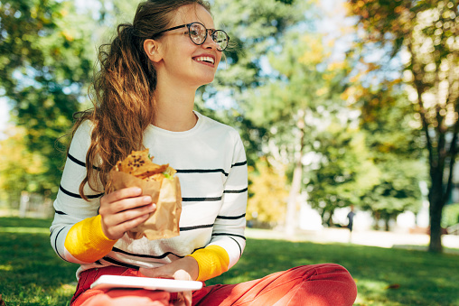 Outdoor shot of a cheerful student female in a casual outfit, eyeglasses, sitting on the green grass have lunch during studying. Positive woman takes a rest eating fast food and learning outside.