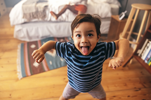 Cute young boy flexing his strength while standing in a bedroom with his sister in the background. Adorable little boy sticking his tongue out playfully.