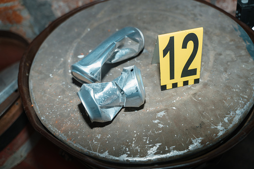 Two beer cans on an oil drum with criminologist's number next to them at the crime scene, we see it illuminated by camera flash