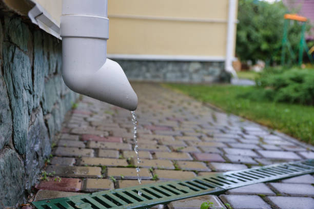 Rainwater flows down a white drainpipe of a beige house into a gutter with a grate, between paving slabs. Beige house with pipes for rain removal.