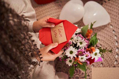 Close up view of the curly woman holding greeting card with I love you signs at her hands while taking it from the bouquet with flowers