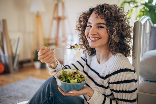 Young woman sitting on the floor at home and eating a salad.