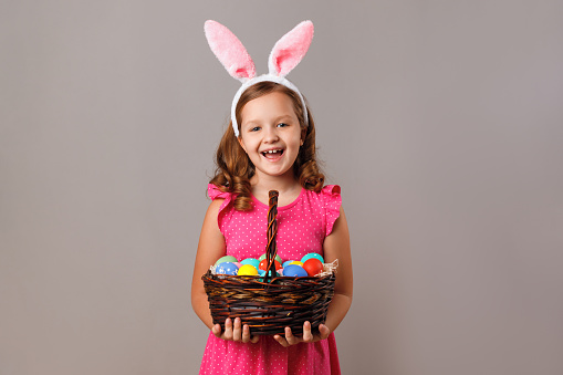 A cute little girl in bunny ears is holding a basket of Easter eggs. Portrait of a child on a gray isolated background.