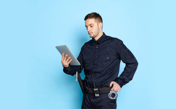 Portrait of young man, policeman officer wearing black uniform using tablet isolated on blue background. Concept of job, caree, law and order. Portrait of young man, policeman officer wearing black uniform using tablet isolated on blue background. Concept of emotions, job, caree, law and order. Security service. Copy space for ad prison guard stock pictures, royalty-free photos & images