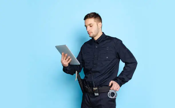 Portrait of young man, policeman officer wearing black uniform using tablet isolated on blue background. Concept of emotions, job, caree, law and order. Security service. Copy space for ad