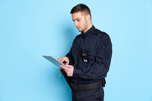 Remote work. Portrait of young man, policeman officer wearing black uniform using tablet isolated on blue background. Concept of emotions, job, caree, law and order. Security service. Copy space for ad