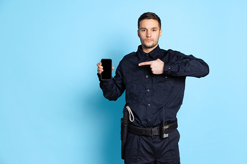 Showing gadget's screen. Portrait of young man, policeman officer wearing black uniform using phone isolated on blue background. Concept of emotions, job, caree, law and order. Security service.