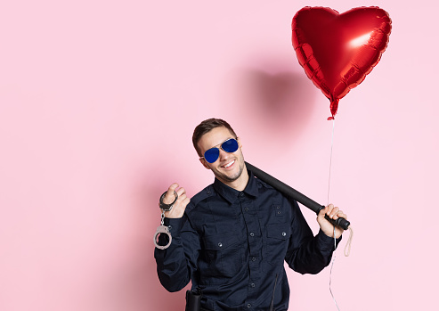 Congrats. Young charming policeman officer with heart shape balloon wearing sunglasses isolated on pink background. St, Valentine's day celebration. Concept of job, safety, holidays, fun and love