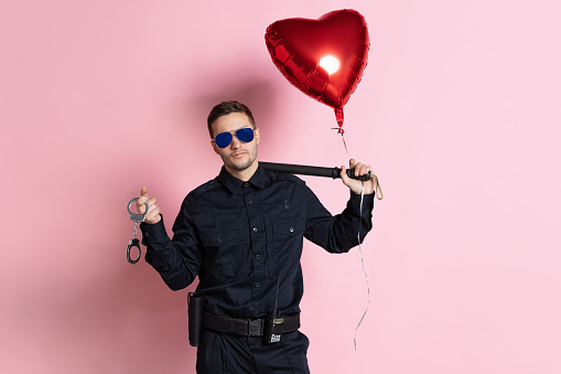 Young charming policeman officer with heart shape balloon wearing sunglasses isolated on pink background. St, Valentine's day celebration. Concept of job, caree, safety, holidays, fun and love