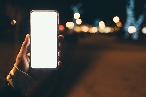 Hand holds smartphone with blank display against background of night city street, close-up