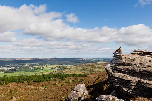 A senior man sitting on a cliff edge and looking at the view while out on a hike in Rothbury, Northumberland. The beautiful green scenery is surrounding him.