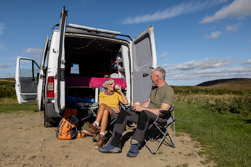 A woman and her son sitting outside the back of their open camper van on camping chairs while on a journey in Rothbury, Northumberland. They are resting after hiking with a cup of tea and are talking with each other.