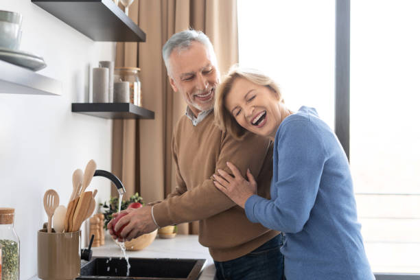 ältere frau lacht, während ihr mann apfel wäscht - two parent family indoors home interior domestic kitchen stock-fotos und bilder