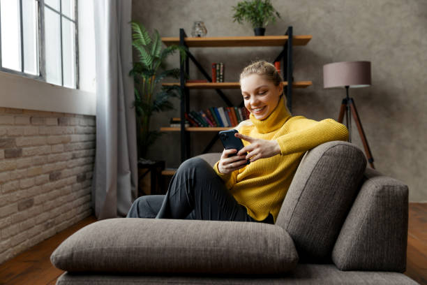 Cheerful young woman using mobile phone while sitting on a sofa at home. stock photo