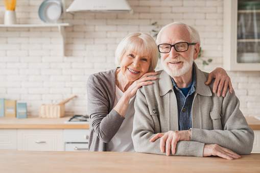 Portrait of caucasian smiling senior old elderly couple family spouses grandparents looking at camera, embracing hugging with love and care at home kitchen