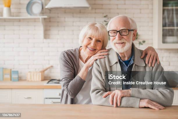 Portrait Of Caucasian Smiling Senior Old Elderly Couple Family Spouses Grandparents Looking At Camera Embracing Hugging With Love And Care At Home Kitchen Stock Photo - Download Image Now