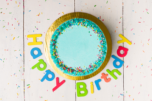 Beautiful delicious cake with blue frosting and sprinkles next to a happy birthday sign on a wooden white table