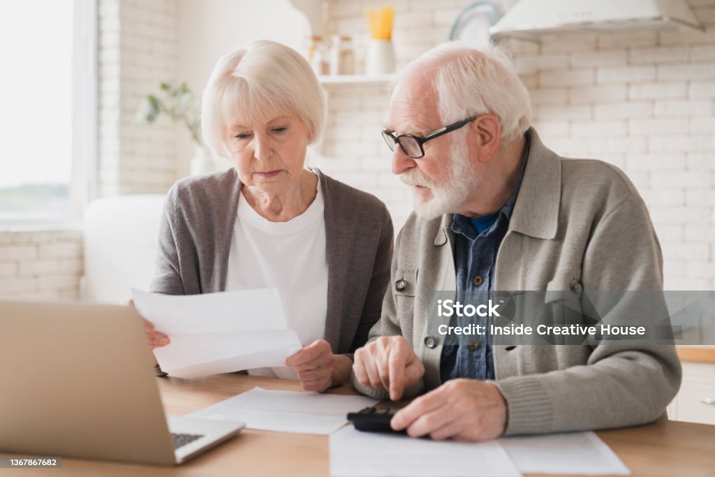 Serious caucasian old elderly senior couple grandparents family counting funds on calculator, doing paperwork, savings, paying domestic bills, mortgage loan, pension at home using laptop. Retirement Stock Photo