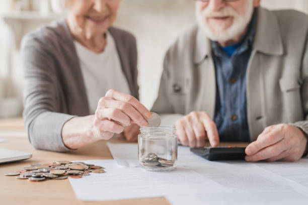 Cropped closeup focused image. Savings concept. Caucasian old elderly senior grandparents couple husband wife spouses putting coin into moneybox, economy for nest egg, pension, mortgage loan at home stock photo