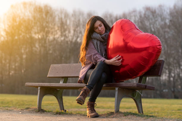 joven triste abrazando un globo en forma de corazón sentado en un banco en el parque - grave nature usa city life fotografías e imágenes de stock