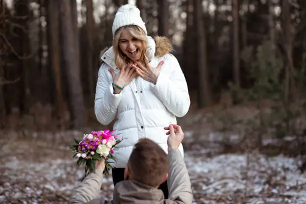 Photo of Portrait of two people couple smiling woman and man proposal ring with bunch of flowers kneeling together outdoors blur