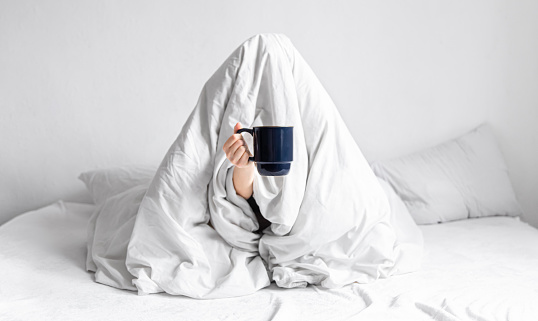 Morning, comfort and people concept - a young woman with a cup of coffee sits in bed under a blanket in a home bedroom.