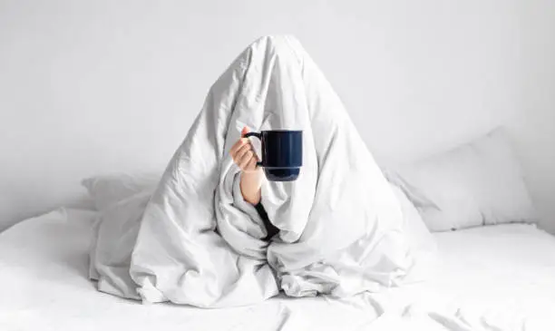 Photo of A woman wrapped in a blanket sits in bed with a cup of coffee in her hands.