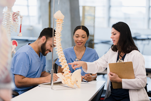 Two young adult students smile and take notes as the mid adult female medical school professor teaches them about the structure of the human spine using a visual aid.