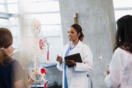 Standing by the model of the human skeleton, the mid adult female medical school professor answers an unseen student's question.