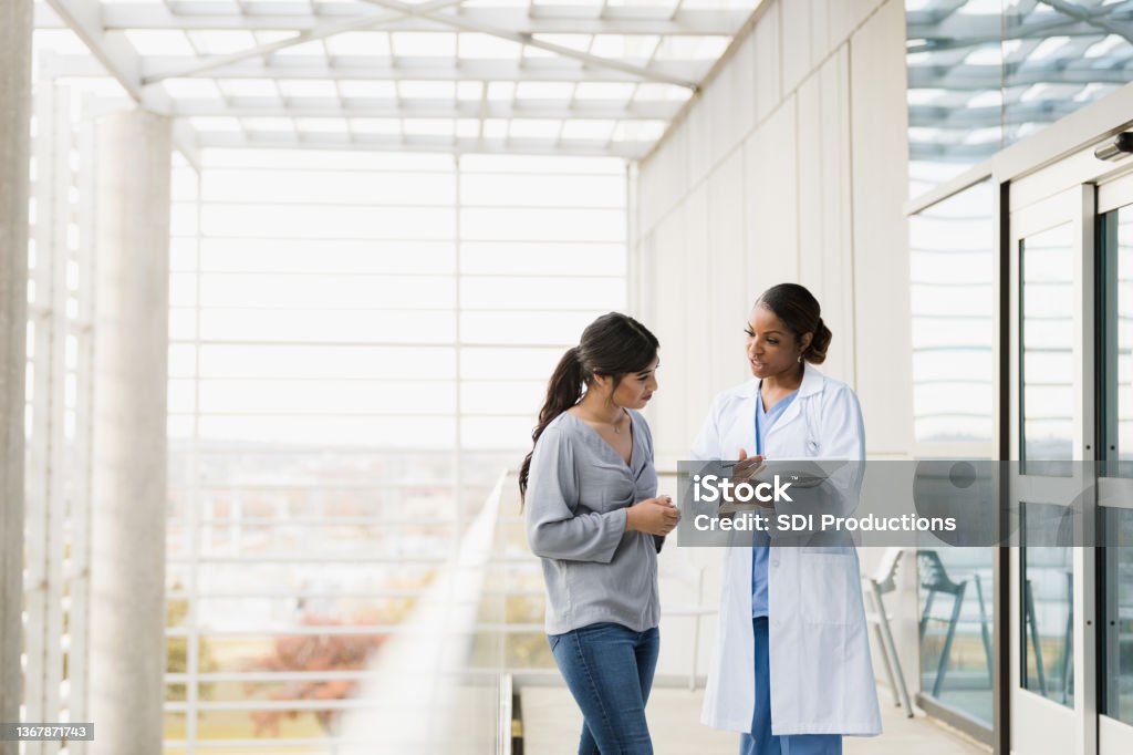 Doctor explains patient's test results to family member The mid adult female doctor explains the patient's test results to the young adult female family member as they stand in the hospital walkway. Doctor Stock Photo