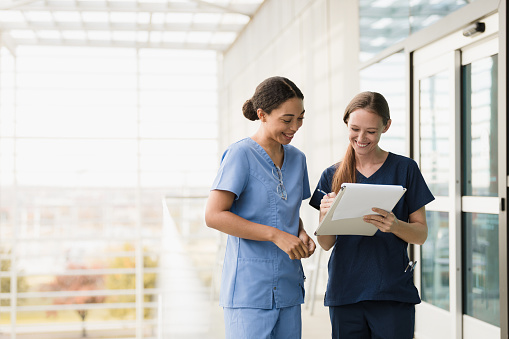 Standing in the walkway of the medical building, the two female nurses smile as they discuss the medical chart.