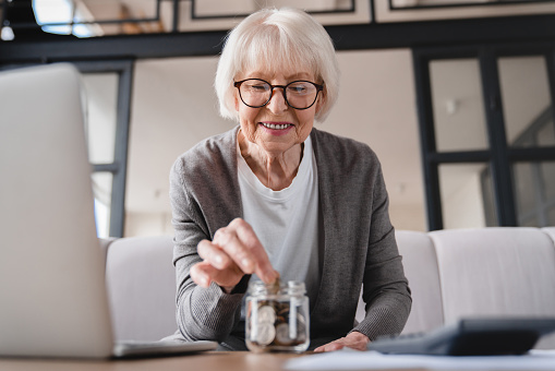 Savings concept. Nest egg of old elderly senior woman grandmother saving money, economizing pension, mortgage loan at home using laptop and putting coin into moneybox