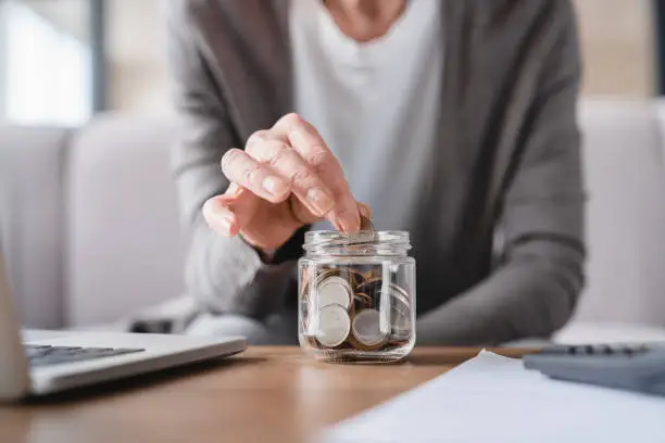 Photo of Cropped focused shot. Savings concept. Nest egg of old elderly senior woman grandmother saving money, economizing pension, mortgage loan at home using laptop and putting coin into moneybox