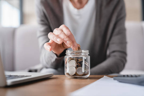 cropped focused shot. savings concept. nest egg of old elderly senior woman grandmother saving money, economizing pension, mortgage loan at home using laptop and putting coin into moneybox - savings finance education mortgage imagens e fotografias de stock