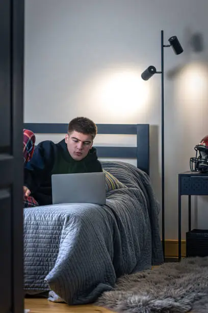 A teenager guy sits in a room on a bed and uses a laptop, a protective sports helmet on the nightstand, the concept of sports.