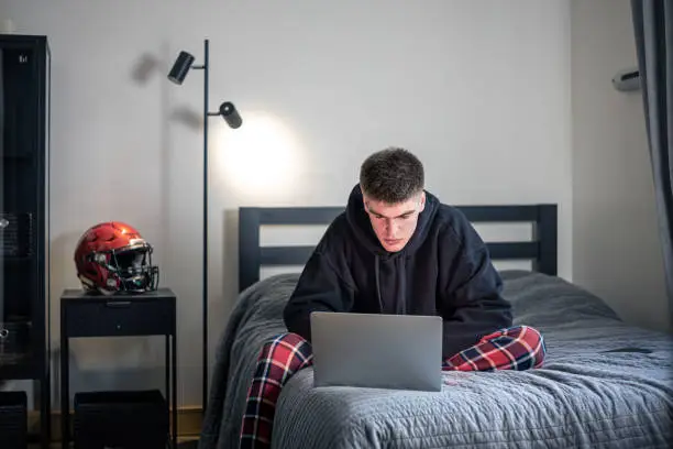 A teenager guy sits in a room on a bed and uses a laptop, a protective sports helmet on the nightstand, the concept of sports.