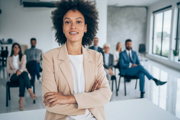 Smiling public speaker in front of her colleagues in conference hall. Shot of confident business leader standing in a board room with crossed arms and looking at camera. Businesswoman leading a training class for professionals. announce stock pictures, royalty-free photos & images