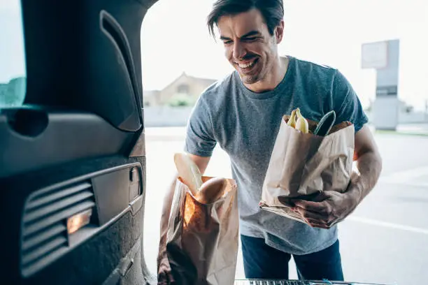 Shot of handsome smiling young man loading paper shopping bags in his car trunk.