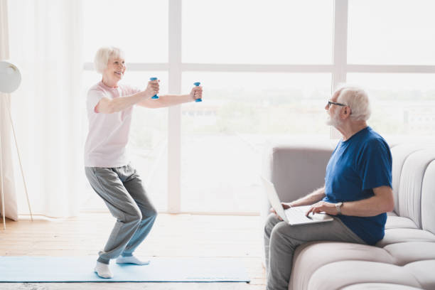 istock Old elderly senior grandparents training together, husband working using laptop while his wife stretching, doing physical exercises with dumbbells at home on lockdown 1367874113