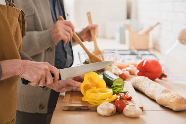 closeup cropped focused image of old senior elderly couple grandparents family spouses cooking together, cutting vegetables at home kitchen, making vegan vegetarian salad - cooking senior adult healthy lifestyle couple imagens e fotografias de stock