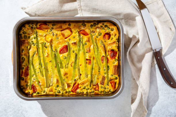 Savory pie with asparagus, cherry tomatoes, green pea and corn. Vegetable clafoutis in baking dish on concrete background. Vegetarian cuisine.
