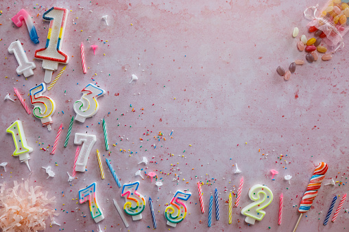 Copy space shot of number shaped birthday candles, jelly beans and colorful sprinkles, lollypop and confetti arranged on a pink background.