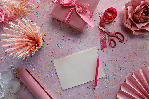 Flat lay of an empty note, a pencil, ribbon, scissors, gift box with a cute bow on top, drinking straws, colorful sprinkles all around and some paper decorations lying on a pastel pink background.