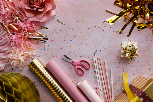 Copy space shot of various wrapping papers, streamers, paper decorations, drinking straws, scissors, gift boxes and colorful sprinkles scattered all around a pastel pink background.