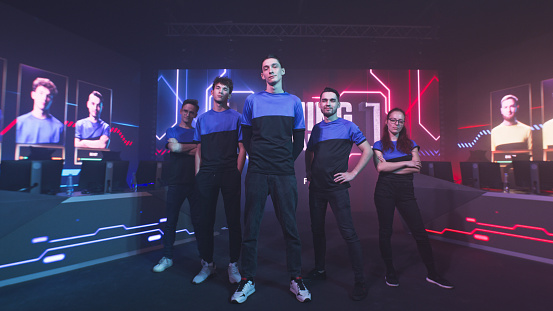 Assured team of esportsman gamers in blue uniform looking at camera while standing under neon illumination during professional gaming championship