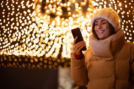 Close up portrait of beautiful smiling woman in white winter hat and jacket while she holds and uses her smart phone during walking outdoors with city christmas illumination on background