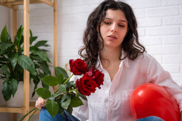 displeased upset girl with bouquet of rose flowers on bed red heart shape balloons. sad, offended, disappointed girl with long hair expressing negative emotions in valentines day stock photo